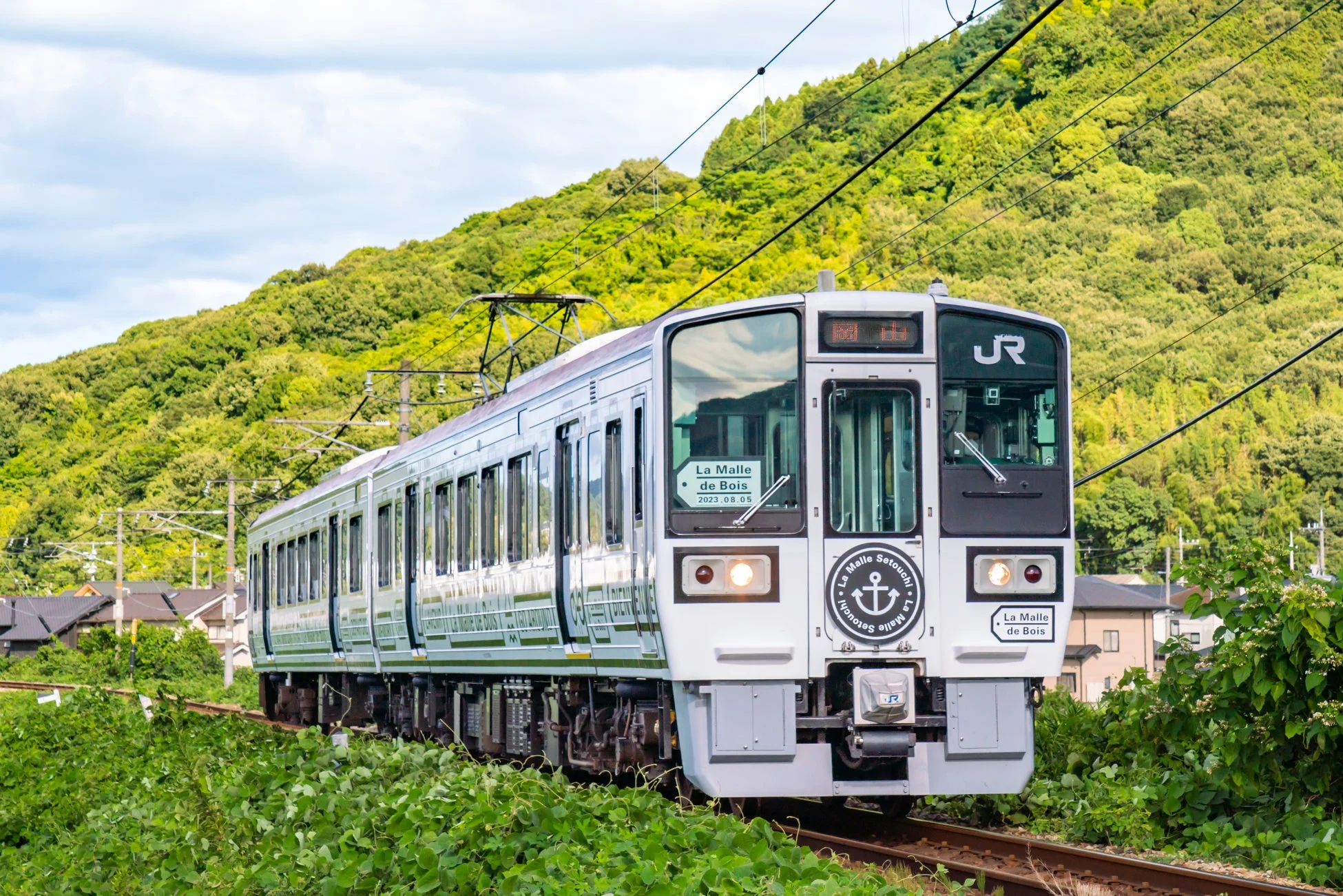 緑豊かな山々を背景に、白い車体に錨のマークが特徴的な観光列車「La Malle de Bois」が線路を走行している