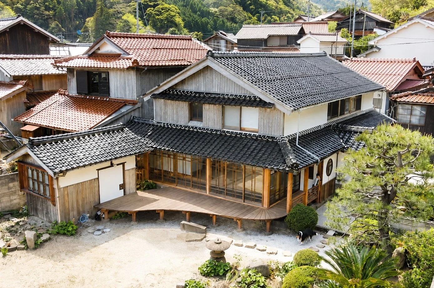 日本の伝統的な家屋が立ち並ぶ集落の風景。手前には大きな縁側を持つ古民家があり、庭には石灯籠や手入れされた木々、そして犬が1匹見えます。山々に囲まれたのどかな日本の里山の日常を捉えた一枚です。