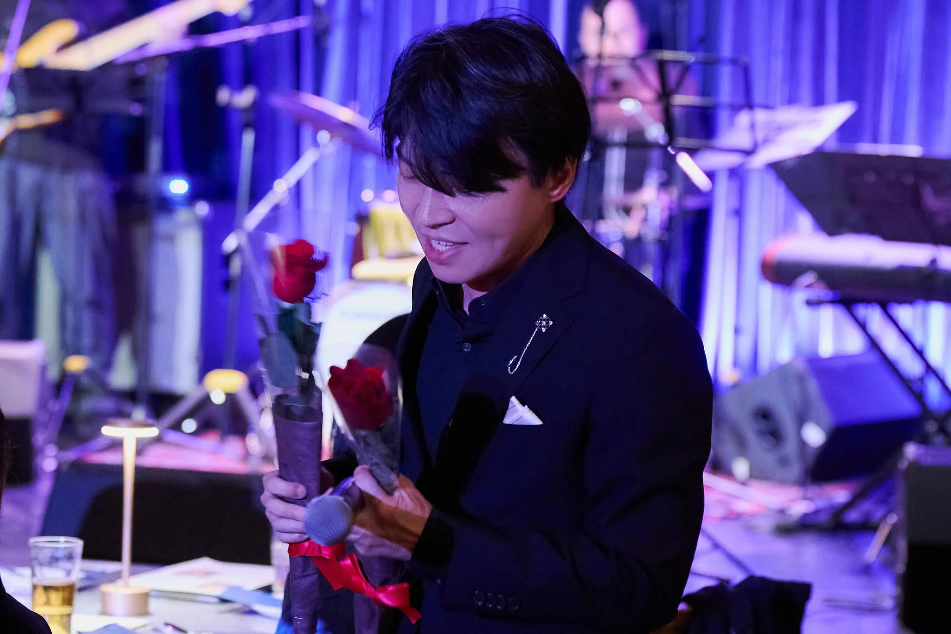 青い照明に照らされたステージで、赤いバラの花束とマイクを持つ川元清史氏