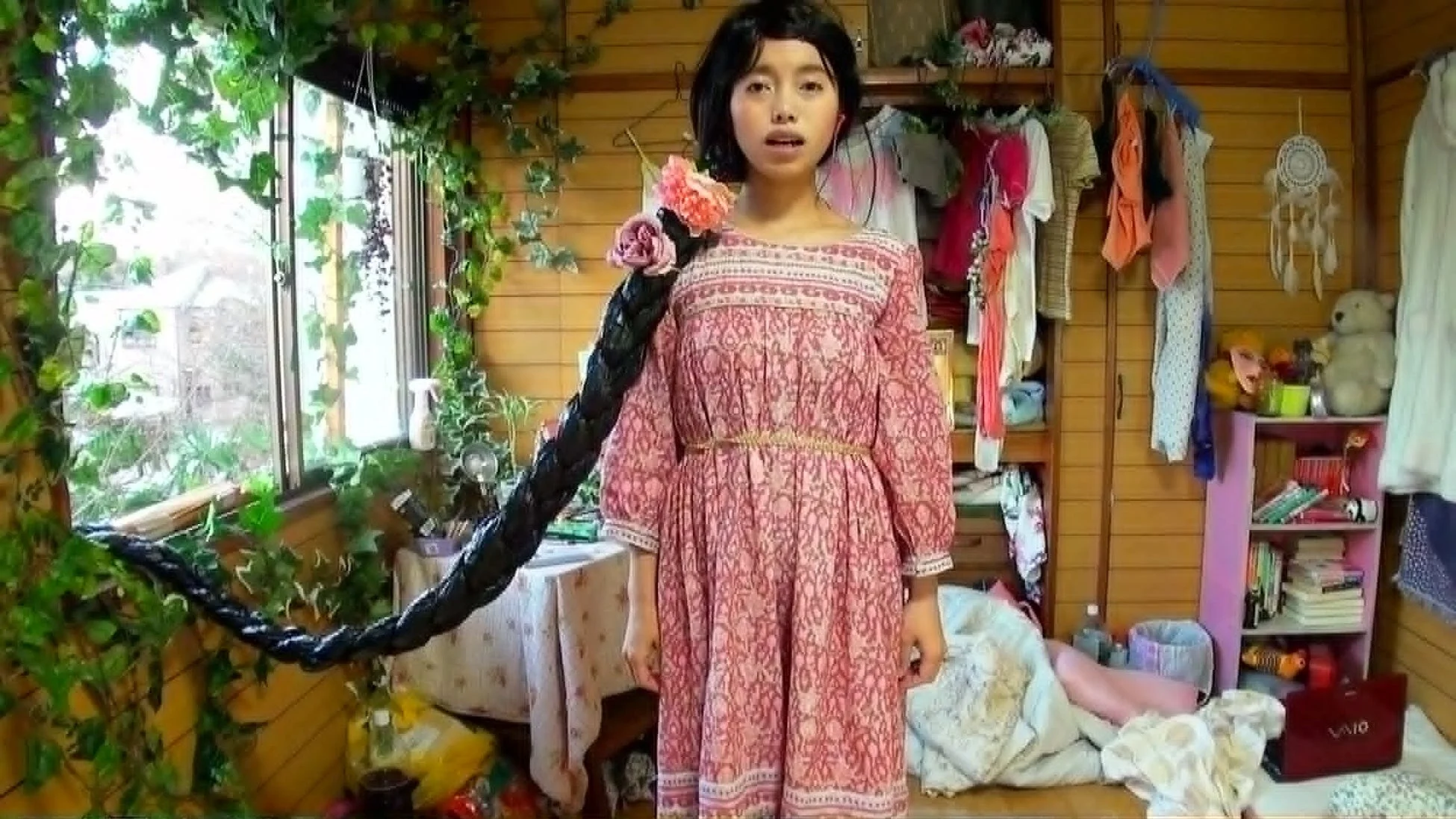つる植物で飾られた窓のある木造の部屋に立つ、長い三つ編みを垂らした少女