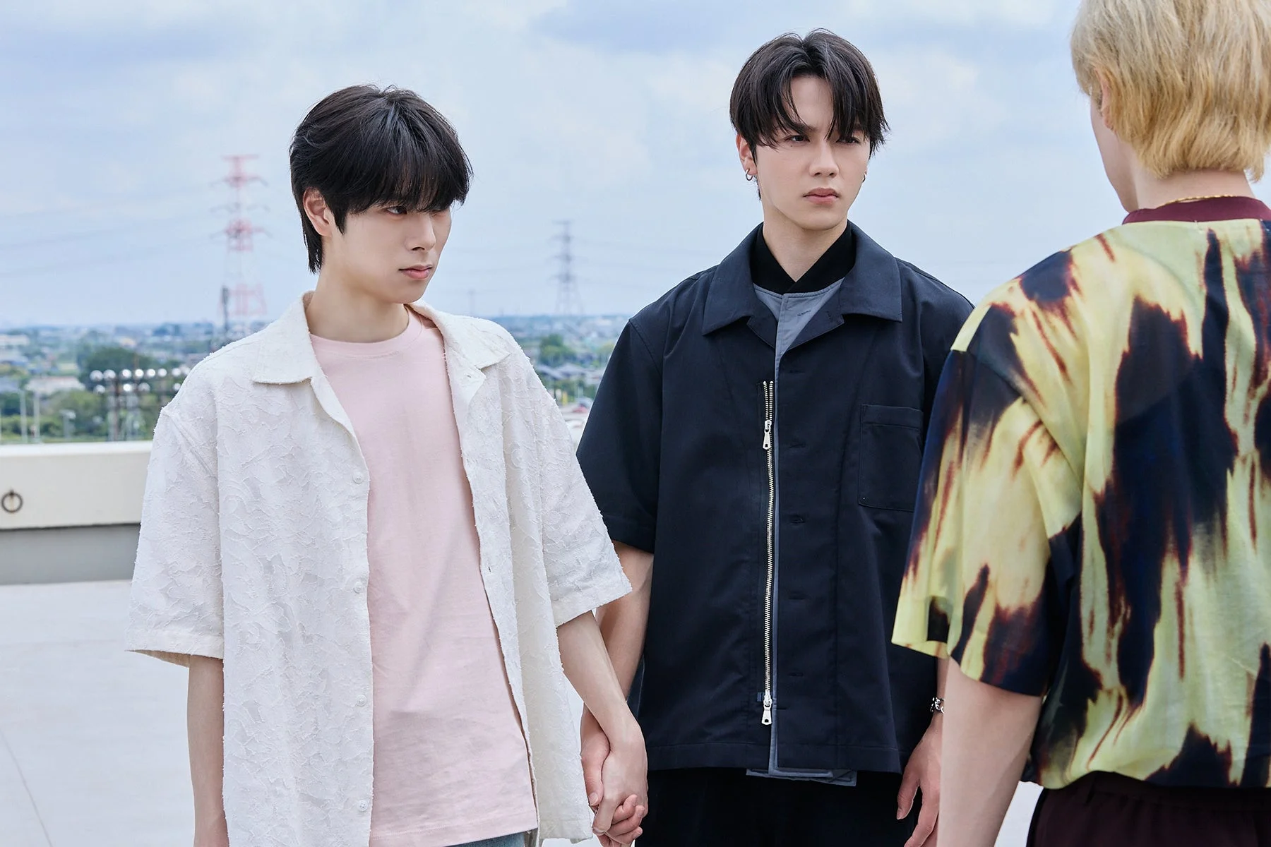屋外で3人の若い男性が並んでおり、左側の2人が手をつないでいる様子。