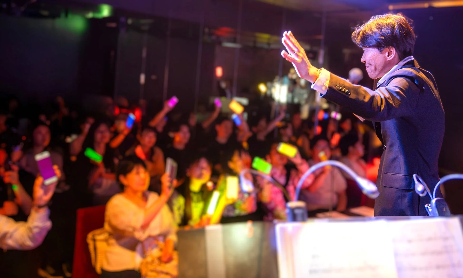 ライブステージで歌唱する男性と、カラフルな光るスマートフォンやペンライトを掲げる観客