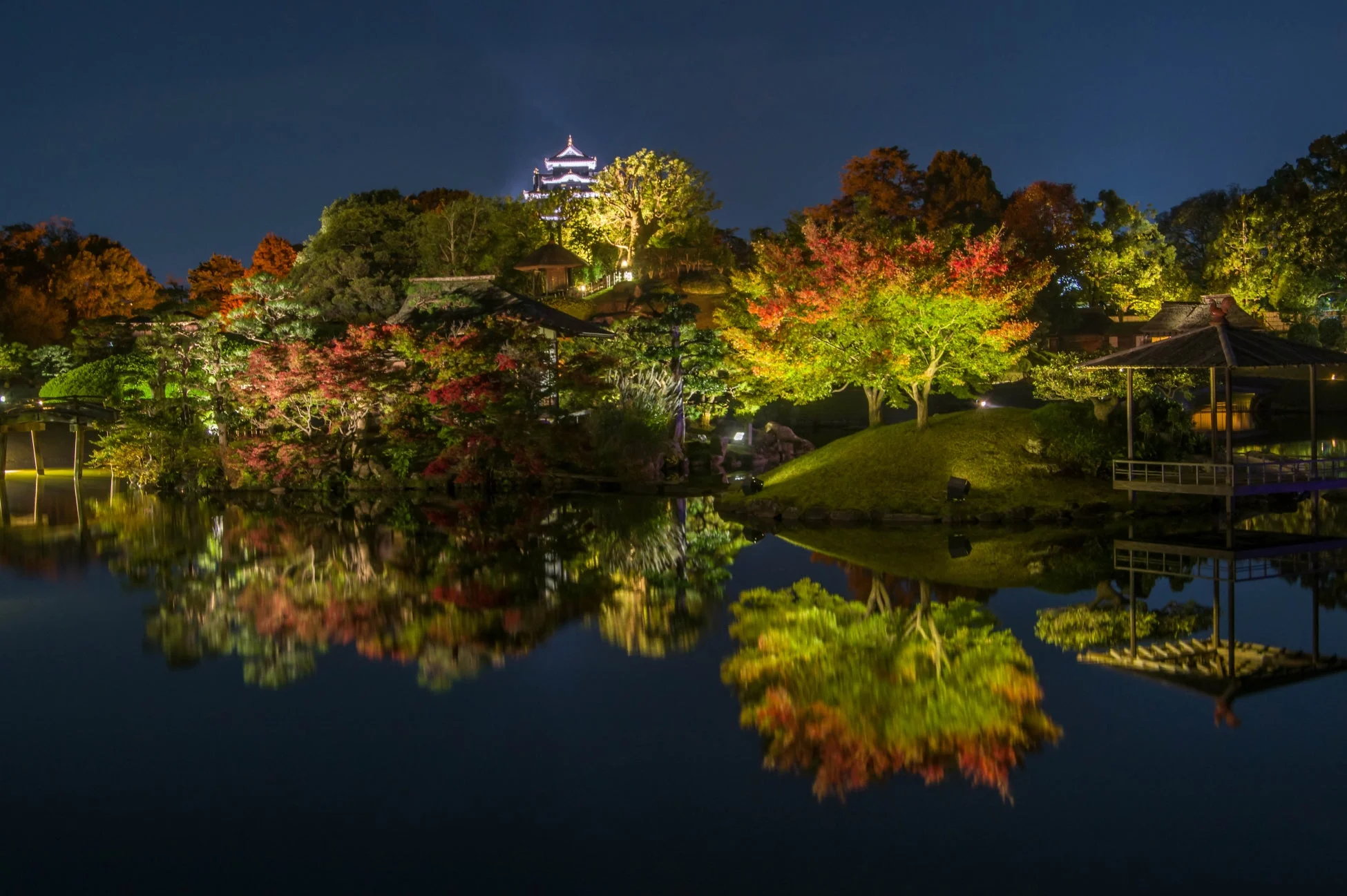 ライトアップされた日本庭園の夜景で、色鮮やかな紅葉の木々や奥に見える城が池に美しく映り込んでいる