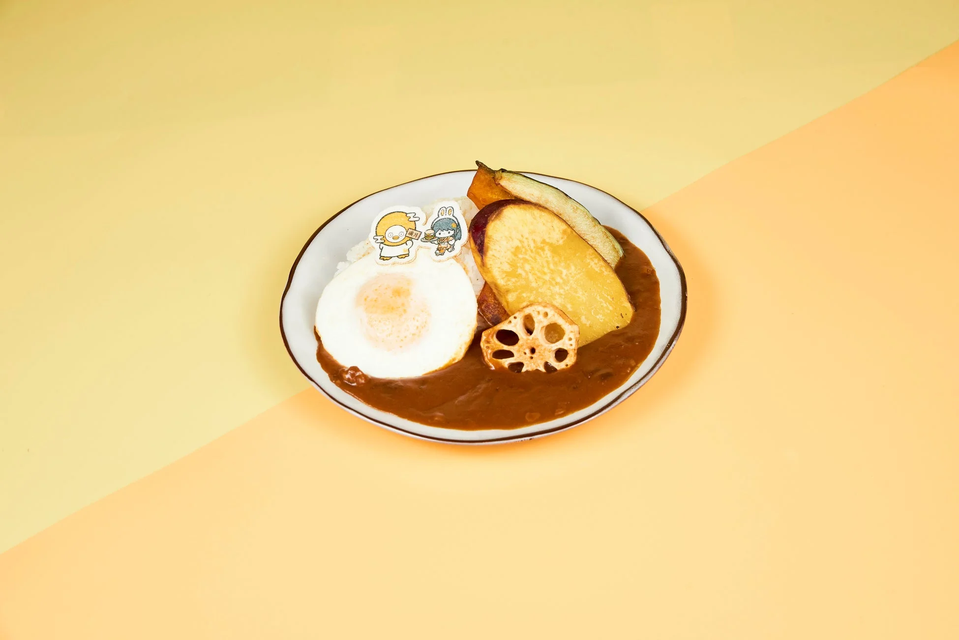 目玉焼きと揚げ野菜(サツマイモ、ズッキーニ、レンコン)がトッピングされたカレーライス。開運と招福の可愛いキャラクターピックが添えられ、明るいツートンカラーの背景に置かれている。
