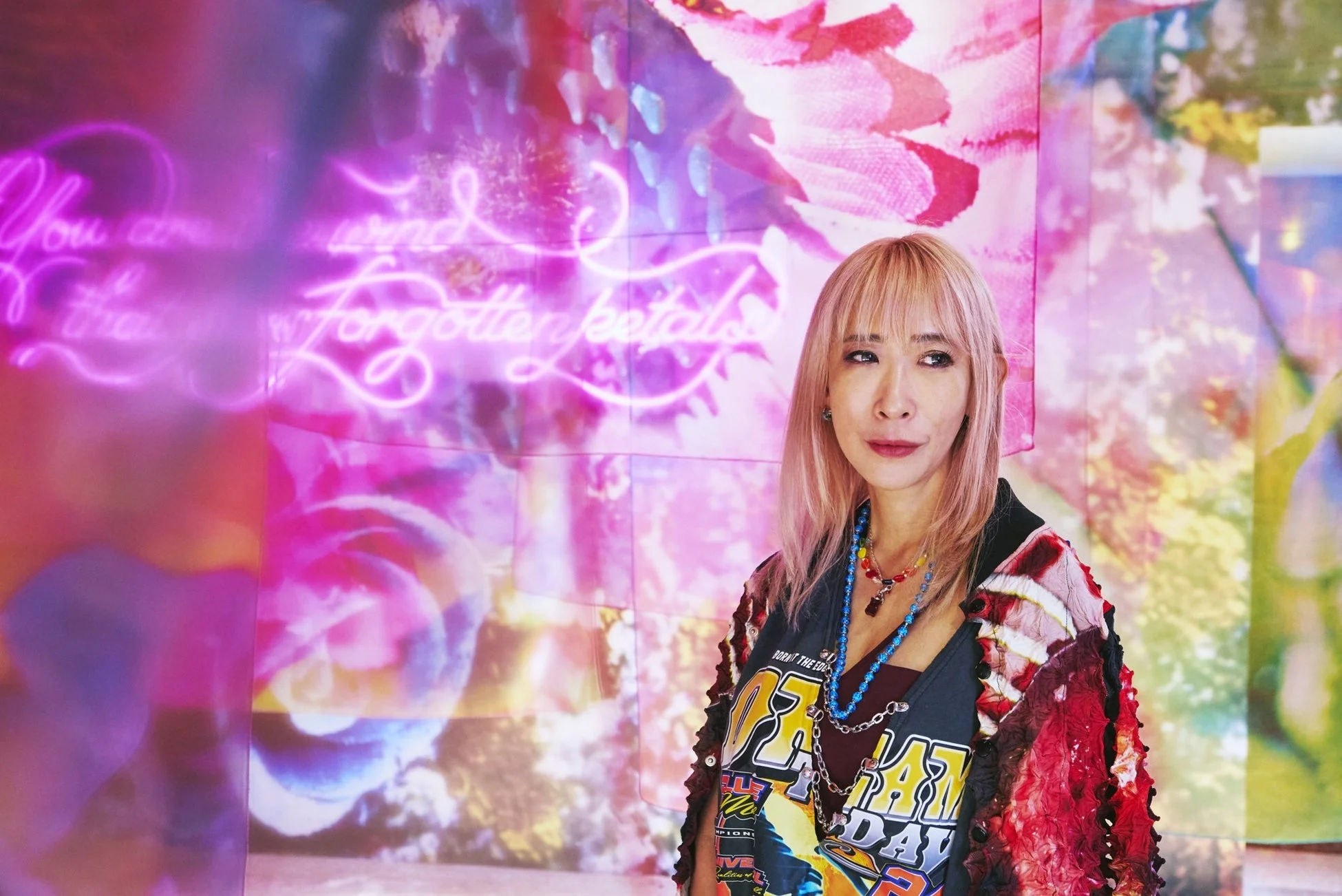 ピンク色の髪と個性的なファッションの女性が、花柄とネオンサインが特徴の鮮やかなアート作品を背景に立っている