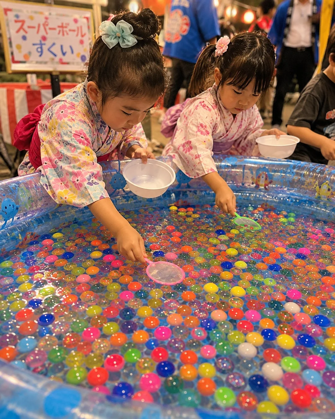 浴衣を着た2人の幼い女の子が、お祭りでスーパーボールすくいを楽しんでいる様子。カラフルなスーパーボールが浮かぶプールから、真剣な表情でボールをすくっている。