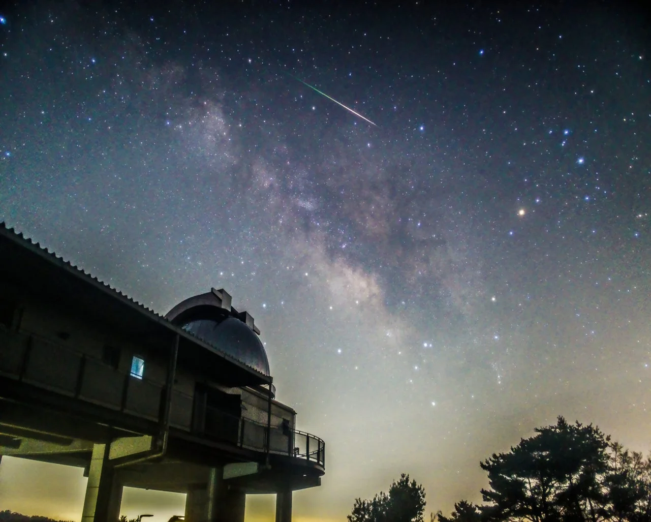 満天の星が輝く夜空に天の川と流星が見え、手前にはドーム型の観測所と木々が写っている