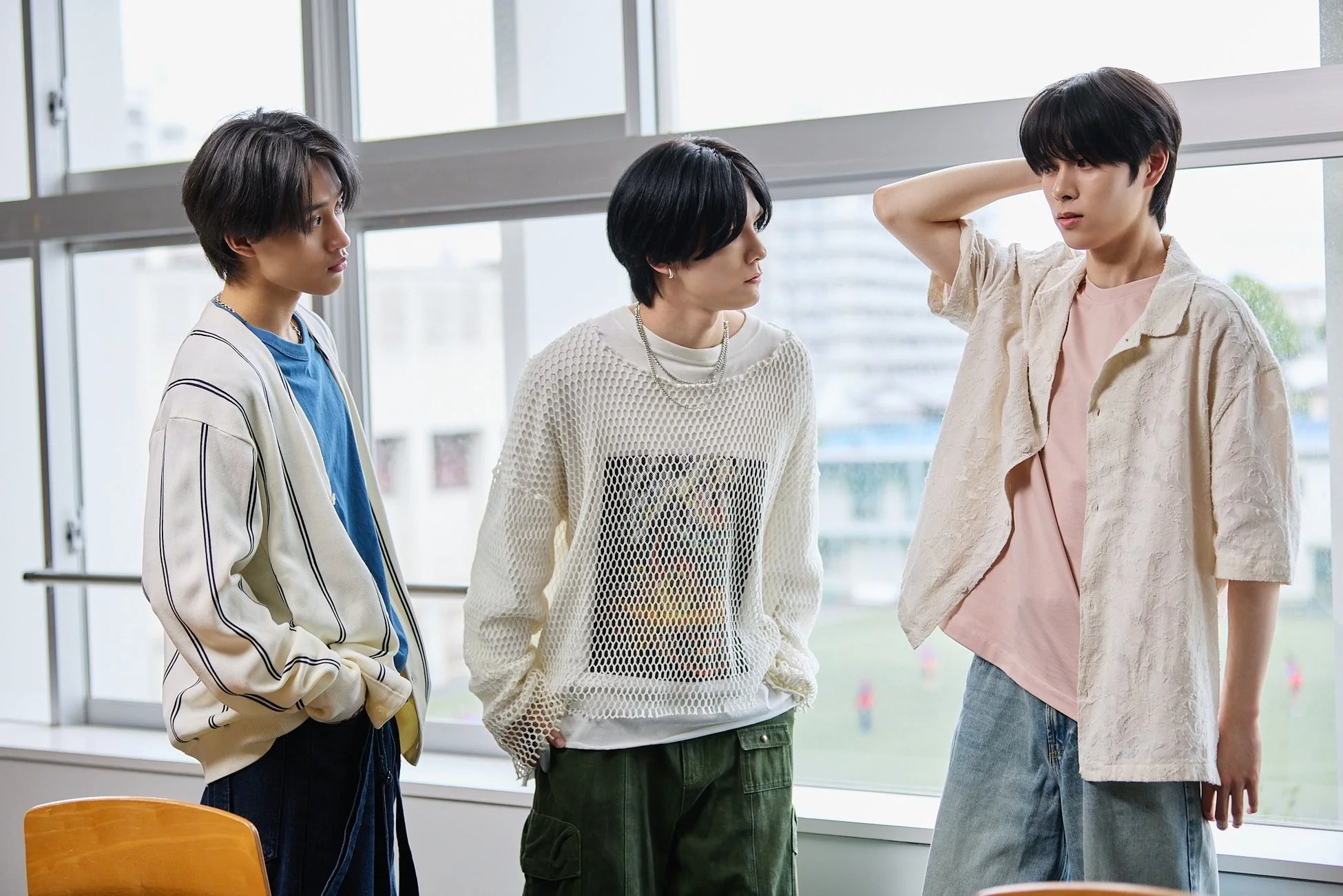 室内で窓際に立つ3人の若者が、それぞれ個性的なカジュアルファッションを着こなしている。